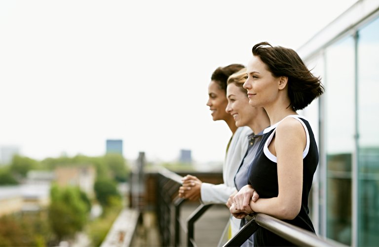 Drei Frauen stehen lächelnd auf einem Balkon mit Blick auf eine grüne Stadtlandschaft. Sie wirken entspannt und optimistisch.