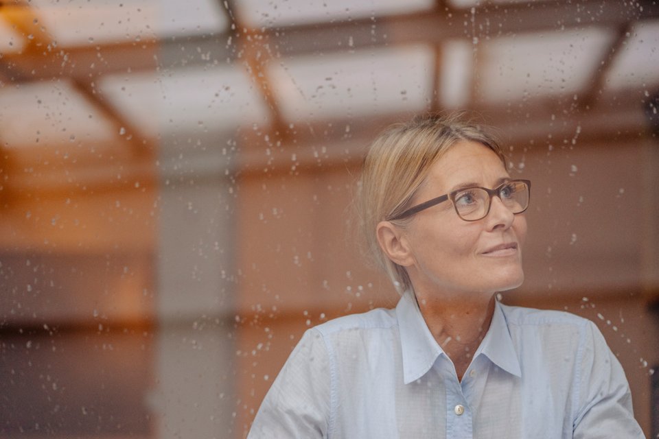 Eine Frau mit blauer Bluse und Brille schaut aus dem Fenster