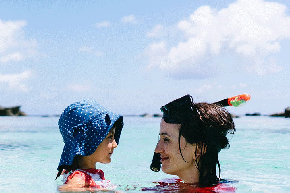 Eine junge Mutter badet mit ihrer Tochter im Meer und beide lächeln fröhlich.