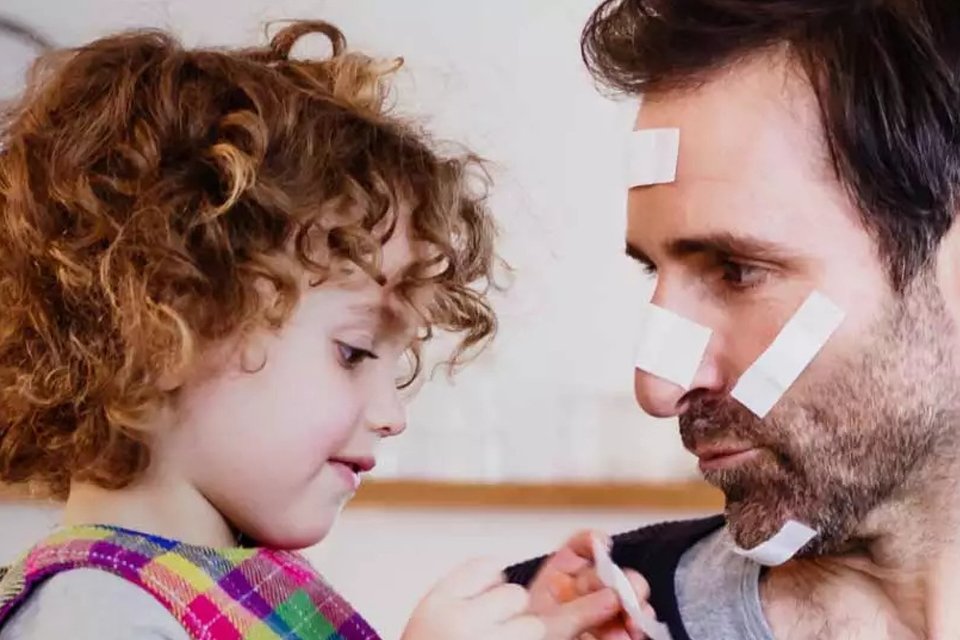 Kleine Tochter mit lockigen Haaren hat Spaß und klebt dem Vater viele Pflaster ins Gesicht.