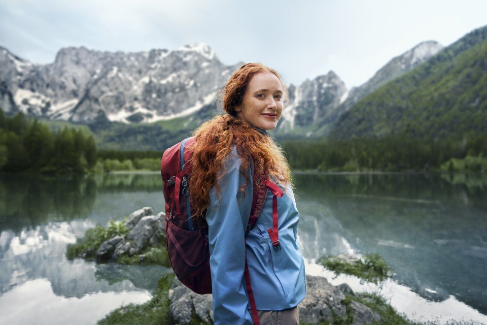 Eine Frau steht vor einem Bergsee und blickt optimistisch in die Kamera