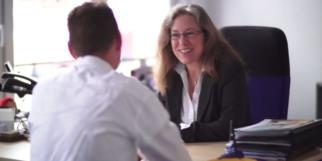 AXA Regionalvertretung Kerstin Zilker in Fürth Eine Beraterin im dunklen Blazer spricht mit einem Kunden im weißen Hemd an einem Schreibtisch. Auf dem Tisch liegen Broschüren und Karten, im Hintergrund ist ein Computerbildschirm mit geöffneter Website zu sehen.