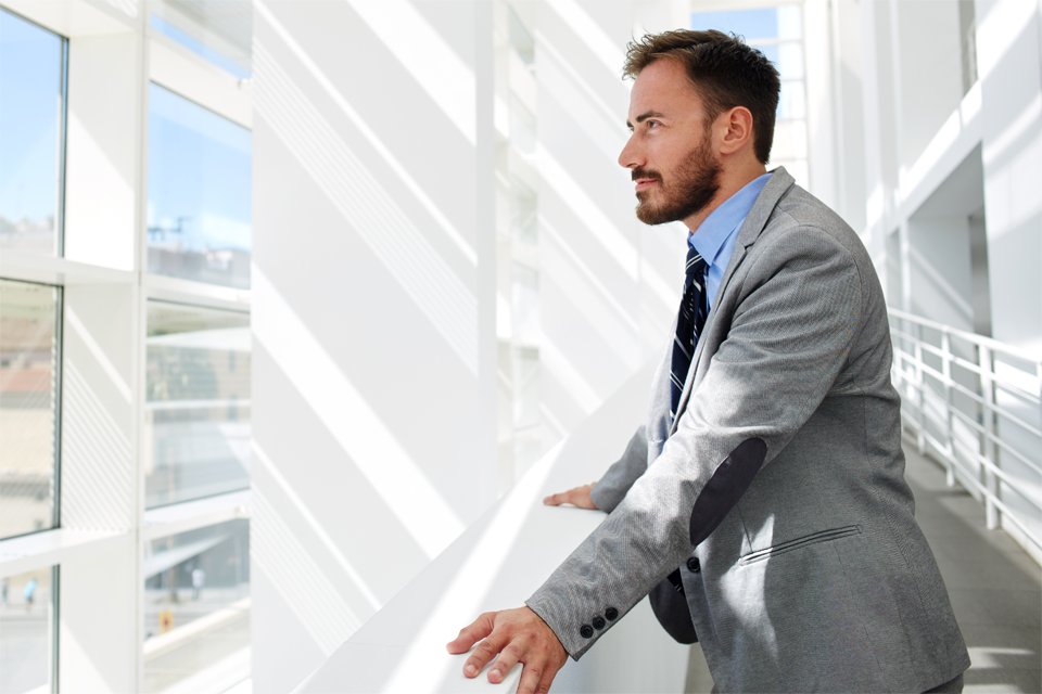 Ein Mann im grauen Anzug mit blauem Hemd und gestreifter Krawatte blickt konzentriert zur Seite. Er befindet sich in einem hellen, modernen Büro mit großen Fenstern und weißen Wänden, durch die Sonnenlicht hereinfällt.